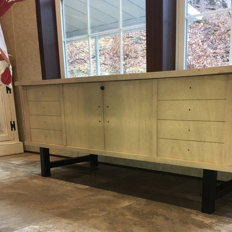 Pu&ograve; includere: Un credenza in legno chiaro con quattro cassetti a sinistra e due ante a destra. La credenza ha gambe in metallo nero e si trova su un pavimento in piastrelle di colore marrone chiaro.