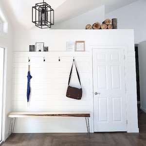May include: A white entryway with a wooden bench, a white door, and a white shiplap wall. There are hooks on the wall with a blue umbrella and a brown leather satchel hanging from them. There are two framed prints on the wall above the hooks.