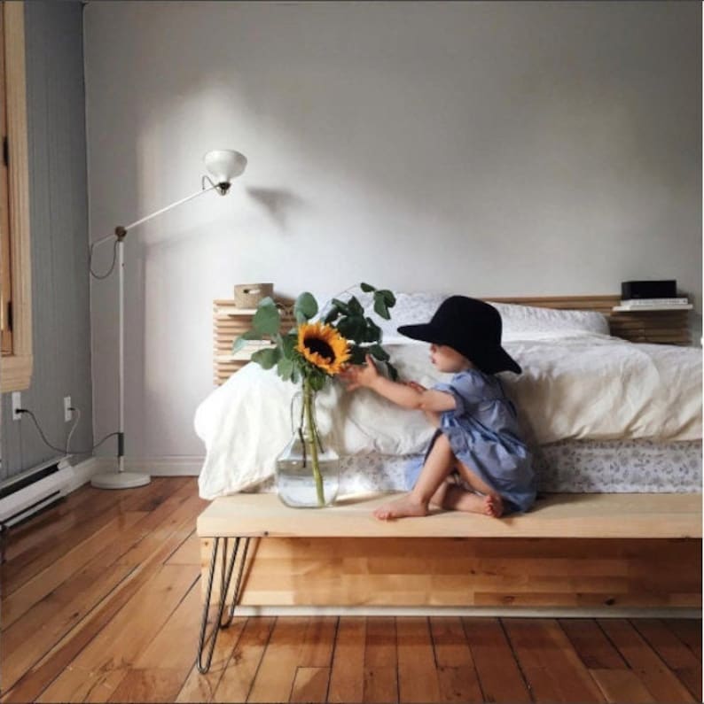 May include: A child wearing a black hat sits on a wooden bench in front of a bed with white linens. A vase with a sunflower and greenery sits on the bench.