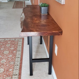 Pu&ograve; includere: Un tavolo console in legno rustico con un piano a bordo vivo e gambe a forcina in metallo nero. Il tavolo &egrave; realizzato in legno riciclato e ha una finitura naturale.