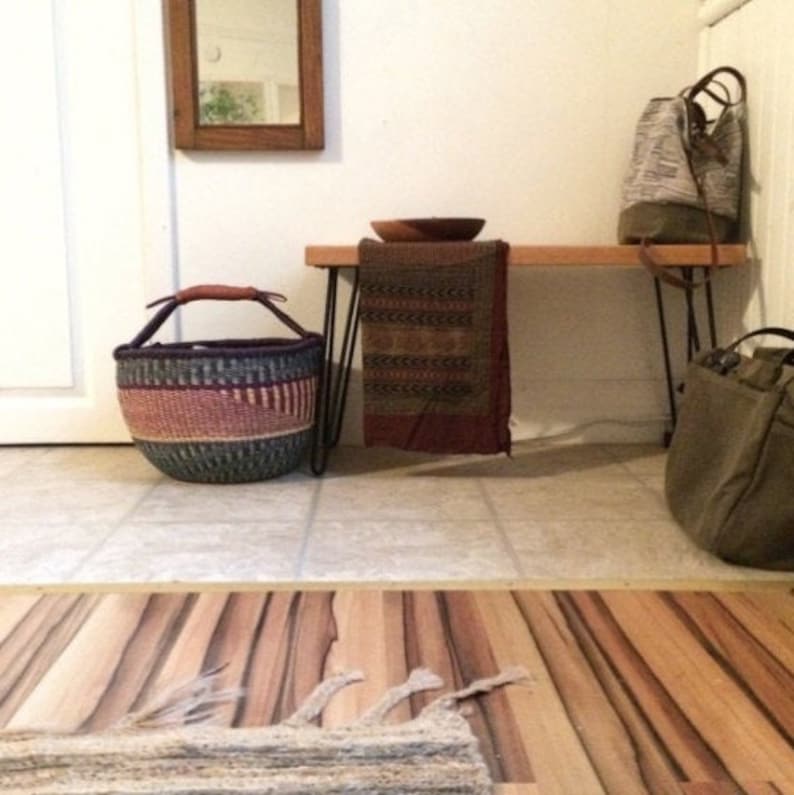 May include: A wooden bench with black hairpin legs, a woven basket, a patterned throw blanket, and two bags. The bench is in a hallway with tiled floors and a wooden floor.