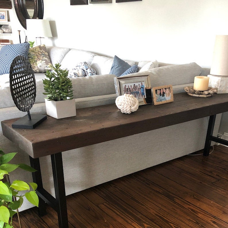 Pu&ograve; includere: Un lungo tavolo console in legno marrone scuro con gambe in metallo nero. Il tavolo &egrave; decorato con una piccola pianta in vaso, un oggetto decorativo in metallo, un nodo di corda bianco e due foto incorniciate.