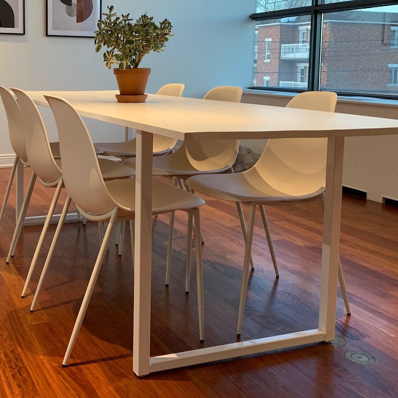 May include: A white rectangular table with a white metal frame and six white chairs. The table is set in a room with hardwood floors and a window in the background.