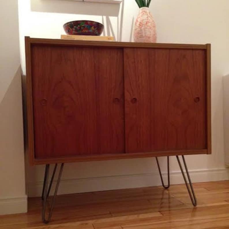 May include: A mid-century modern cabinet with two wooden doors and hairpin legs. The cabinet is made of wood with a rich brown finish.