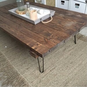 May include: A dark brown wooden coffee table with a rustic finish. The table has a rectangular top and four hairpin legs. The table is sitting on a light brown rug.