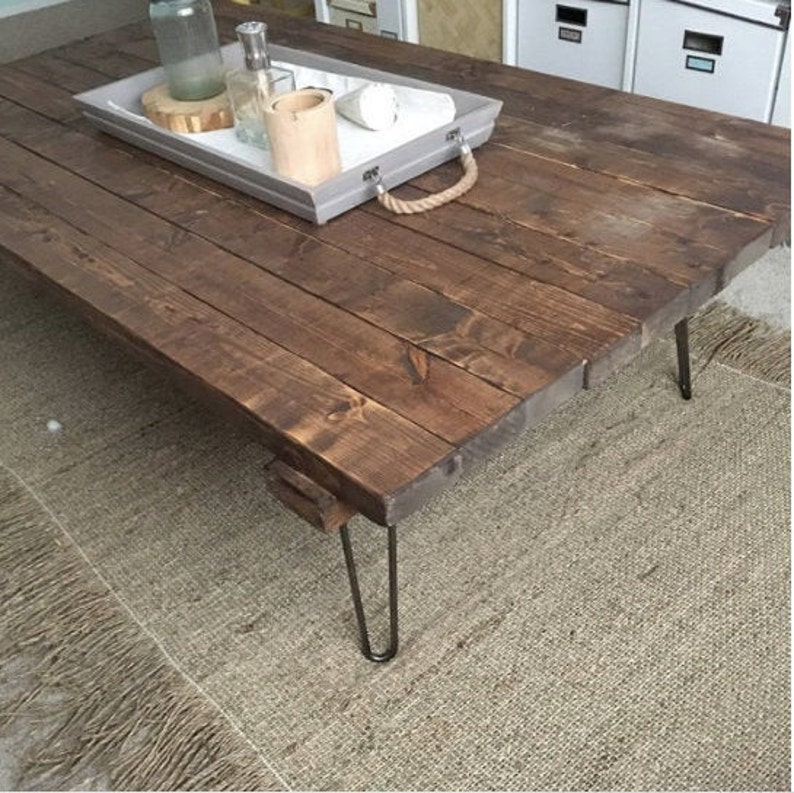 May include: A dark brown wooden coffee table with a rustic finish. The table has a rectangular top and four hairpin legs. The table is sitting on a light brown rug.