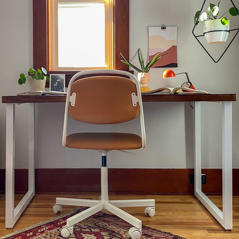 May include: A white desk with a brown wooden top and a white metal frame. A brown office chair with a white base and wheels is positioned in front of the desk. A small potted plant is on the desk, along with a framed photo and a book.