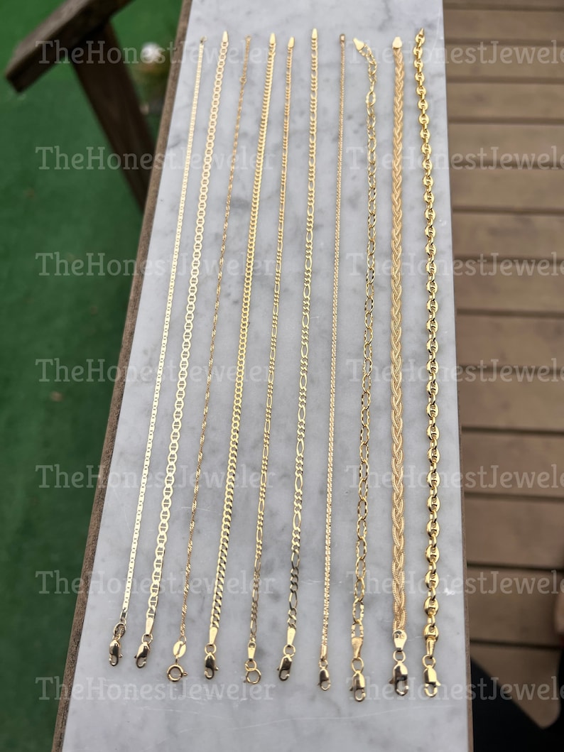 May include: Nine gold chains of different styles and lengths on a white surface. The chains are all made of gold and have a variety of different links.