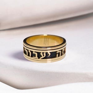 May include: A gold-toned band ring featuring a central cutout design with Hebrew characters in black relief. The ring has a smooth, polished finish on the top and bottom edges, creating a layered appearance with the dark lettering set against the metal.