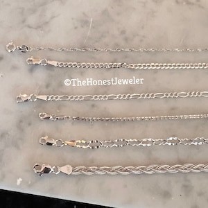 May include: A collection of six silver chains of different styles and lengths, laid out on a white marble surface.  The chains are all made of silver and have a polished finish.  The chains are all different lengths and have different styles of links.  The chains are all laid out in a straight line, with the shortest chain at the top and the longest chain at the bottom.