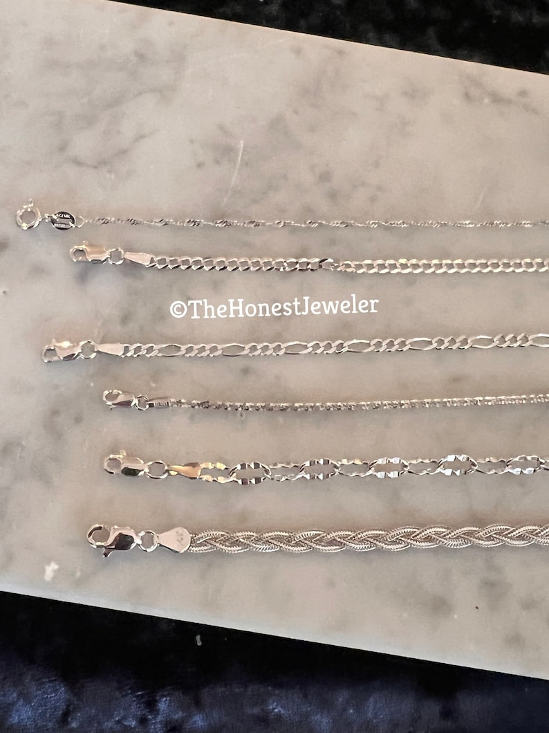 May include: A collection of six silver chains of different styles and lengths, laid out on a white marble surface.  The chains are all made of silver and have a polished finish.  The chains are all different lengths and have different styles of links.  The chains are all laid out in a straight line, with the shortest chain at the top and the longest chain at the bottom.