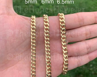 May include: Three gold chains of different widths, 5mm, 6mm, and 6.5mm, are displayed on a hand. The chains are Cuban link style.