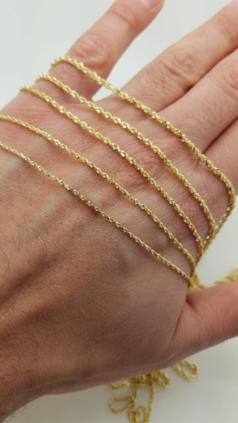 May include: A close-up of a hand holding a variety of gold rope chains. The chains are arranged in order of thickness, with the thinnest chain at the top and the thickest chain at the bottom.