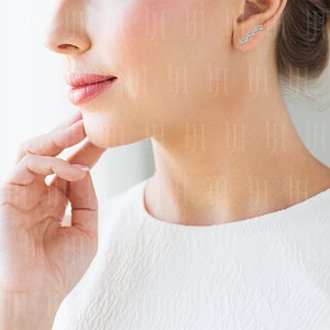 May include: A close-up of a woman's ear with a pair of small, silver earrings featuring a row of three baguette-shaped stones.
