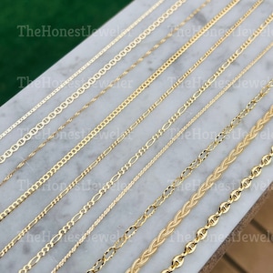 May include: A collection of gold chains of different styles and thicknesses, laid out on a white marble surface. The chains are all made of gold and have a shiny finish.