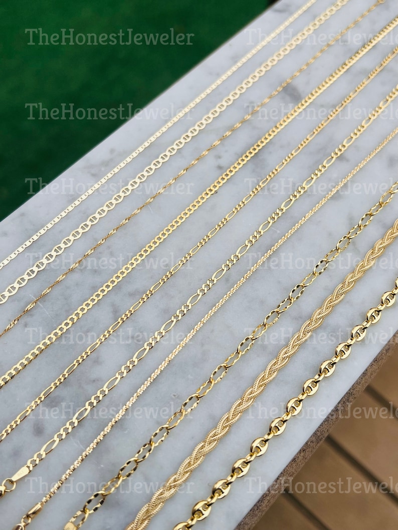May include: A collection of gold chains of different styles and thicknesses, laid out on a white marble surface. The chains are all made of gold and have a shiny finish.