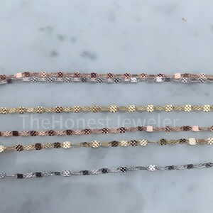 May include: Four chains of different colors, silver, gold, and rose gold, are laid out on a white surface. The chains are made of small, oval-shaped links with a textured surface.