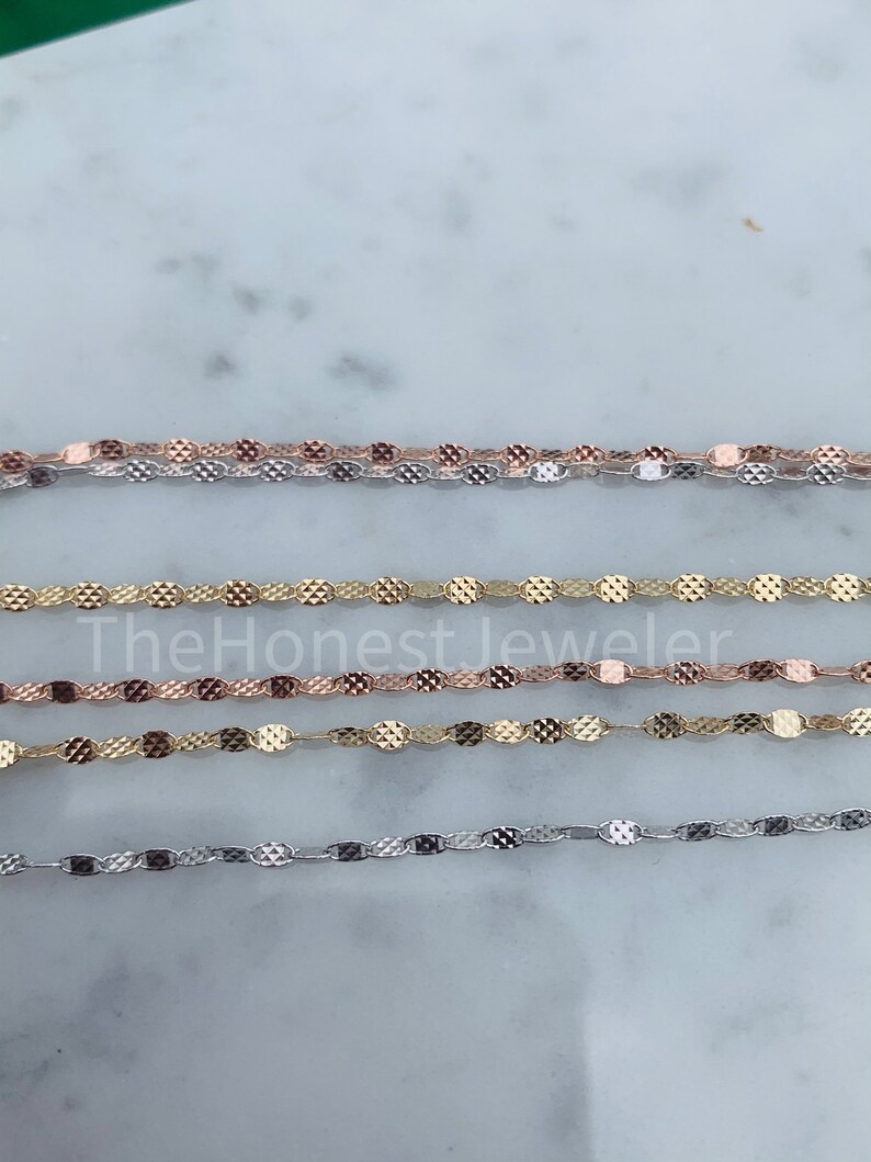 May include: Four chains of different colors, silver, gold, and rose gold, are laid out on a white surface. The chains are made of small, oval-shaped links with a textured surface.