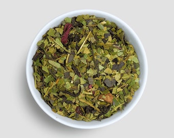 May include: A white bowl filled with a blend of green, black, and red tea leaves. The tea leaves are a mix of loose leaf green tea, black tea, and dried fruit pieces.