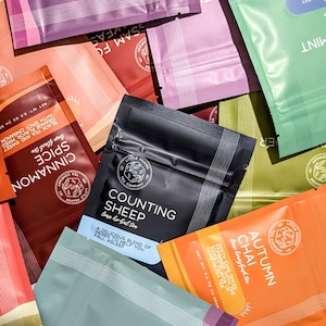 May include: Assortment of tea packets in various colors, including black, orange, green, and pink. The packets feature text such as "Counting Sheep," "Autumn Chai," and "Cinnamon Spice." Each packet is labeled with the "Little Prayer Tea Company" logo.