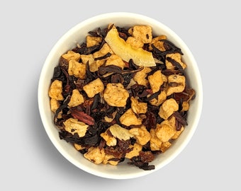 May include: A white bowl containing a loose-leaf tea blend. The tea includes dark red and black tea leaves, alongside pieces of dried fruit in yellow and brown hues. A large, thin slice of dried fruit is placed on top.