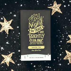 May include: A black box of loose leaf tea with gold lettering that reads "Holy Night the Stars are Brightly Shining" and "Winter Tea" with a small image of a pine tree on each side. The box is on a black background with gold glitter stars.