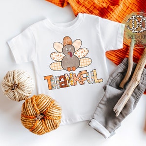 Puede incluir: Camiseta blanca con un dise&ntilde;o de pavo de patchwork y la palabra "Thankful" en letras de colores. La camiseta est&aacute; sobre un fondo blanco con una rama marr&oacute;n, un pantal&oacute;n gris y una calabaza tejida.