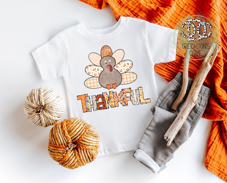 Puede incluir: Camiseta blanca con un dise&ntilde;o de pavo de patchwork y la palabra "Thankful" en letras de colores. La camiseta est&aacute; sobre un fondo blanco con una rama marr&oacute;n, un pantal&oacute;n gris y una calabaza tejida.