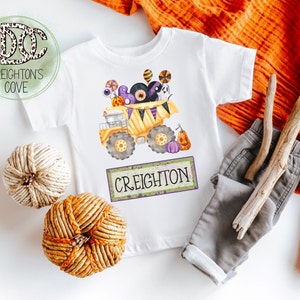 Op de afbeelding: Wit T-shirt met een Halloween-thema van een gele dumper gevuld met snoep, ogen en een spook. De naam "Creighton" staat eronder gedrukt. Het shirt wordt gecombineerd met een grijze broek en decoratieve elementen.