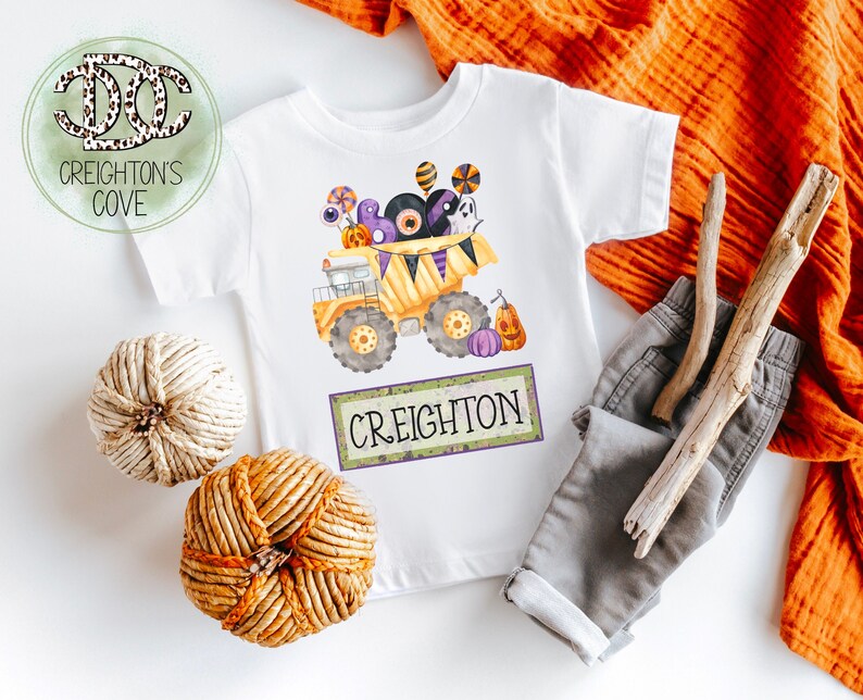 Op de afbeelding: Wit T-shirt met een Halloween-thema van een gele dumper gevuld met snoep, ogen en een spook. De naam "Creighton" staat eronder gedrukt. Het shirt wordt gecombineerd met een grijze broek en decoratieve elementen.