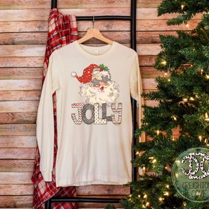 May include: A long-sleeved, cream-colored t-shirt with a festive design featuring Santa Claus wearing sunglasses and the word "Jolly" in colorful letters.