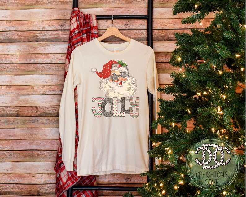 May include: A long-sleeved, cream-colored t-shirt with a festive design featuring Santa Claus wearing sunglasses and the word "Jolly" in colorful letters.