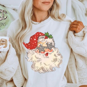 May include: White sweatshirt with a vintage-style illustration of Santa Claus wearing sparkly star-shaped sunglasses. The illustration is in red, white, and green.