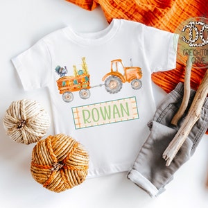 May include: A white baby onesie with a watercolor illustration of a tractor pulling a wagon full of pumpkins and a turkey. The onesie has the name "ROWAN" printed on it.
