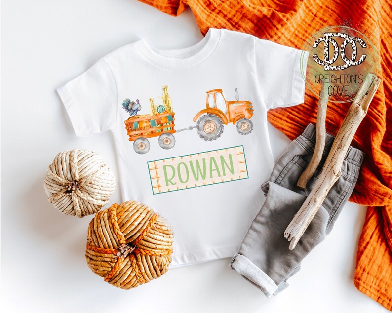 May include: A white baby onesie with a watercolor illustration of a tractor pulling a wagon full of pumpkins and a turkey. The onesie has the name "ROWAN" printed on it.