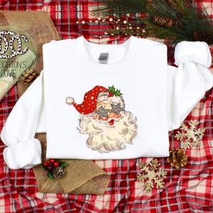 May include: White sweatshirt with a festive design of Santa Claus wearing sparkly star-shaped sunglasses. The design is in a vintage style with red, white, and green colors.