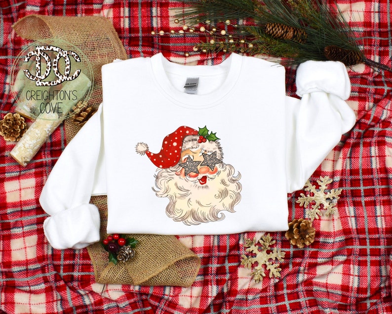 May include: White sweatshirt with a festive design of Santa Claus wearing sparkly star-shaped sunglasses. The design is in a vintage style with red, white, and green colors.