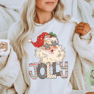 May include: White sweatshirt with a festive design featuring a cartoon Santa Claus wearing sunglasses and the word "Jolly" in colorful patterned letters.