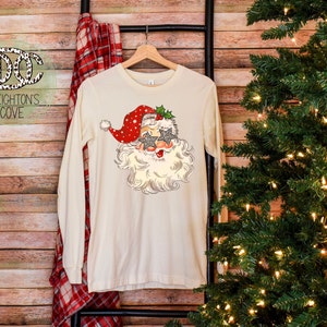 May include: A white long-sleeved t-shirt with a vintage-style illustration of Santa Claus wearing sunglasses and a red hat with white polka dots. The design includes holly and a green bow.