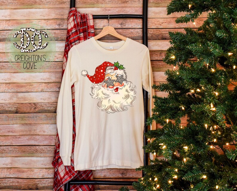 May include: A white long-sleeved t-shirt with a vintage-style illustration of Santa Claus wearing sunglasses and a red hat with white polka dots. The design includes holly and a green bow.