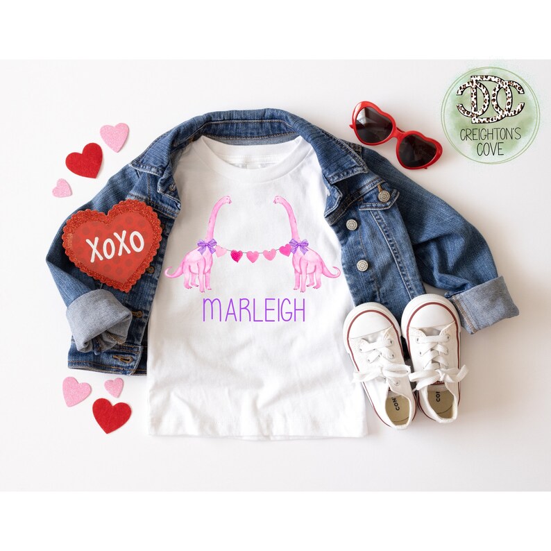 K&ouml;nnte beinhalten: Wei&szlig;es T-Shirt mit einem Paar rosa Dinosaurier, die eine Girlande aus rosa Herzen halten. Das Shirt tr&auml;gt den Namen "MARLEIGH" in lila unter den Dinosauriern.