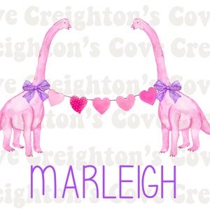 K&ouml;nnte beinhalten: Zwei rosa Dinosaurier mit lila Schleifen halten eine Girlande aus rosa Herzen mit wei&szlig;en Punkten. Der Text "MARLEIGH" ist in lila unter den Dinosauriern geschrieben.