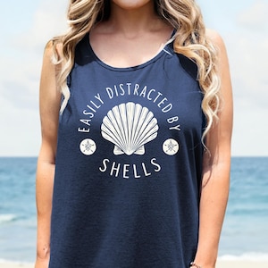 May include: Navy blue tank top with the text "EASILY DISTRACTED BY SHELLS" in white. The design includes a seashell and two starfish. The top is sleeveless and has a scoop neck.