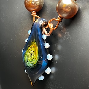 May include: A handcrafted necklace featuring a teardrop-shaped pendant with a swirling yellow and green design within a dark blue glass. The pendant is accented with small, white beads and connected to a copper chain with hammered metal beads.