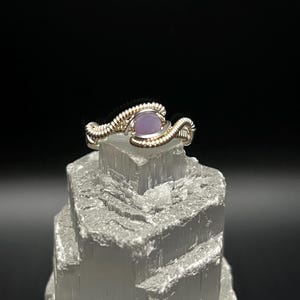 Op de afbeelding: Handgemaakte zilverkleurige ring met een lichtpaarse ovale edelsteen. De ring heeft een draadgewikkeld ontwerp, met de edelsteen in het midden gezet. De ring wordt gepresenteerd op een witte kristalformatie tegen een zwarte achtergrond.
