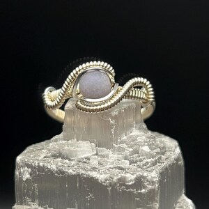 Peut inclure: Une bague en fil d'argent avec une pierre précieuse violet clair au centre. La bague est présentée sur une surface cristalline blanche sur fond noir. Le fil a un design torsadé et texturé.