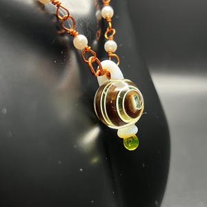 May include: A handcrafted necklace featuring a pendant with a brown and green spiral design. The pendant is suspended from a chain of copper wire and white pearls. A small, green, teardrop-shaped bead hangs below the pendant.