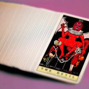 May include: The Devil tarot card features a red, sparkly figure with horns and wings, sitting on a throne. Two figures are chained to the throne, one in red and one in black. The card reads "THE DEVIL ENJOY  YOUR  MISTAKE".