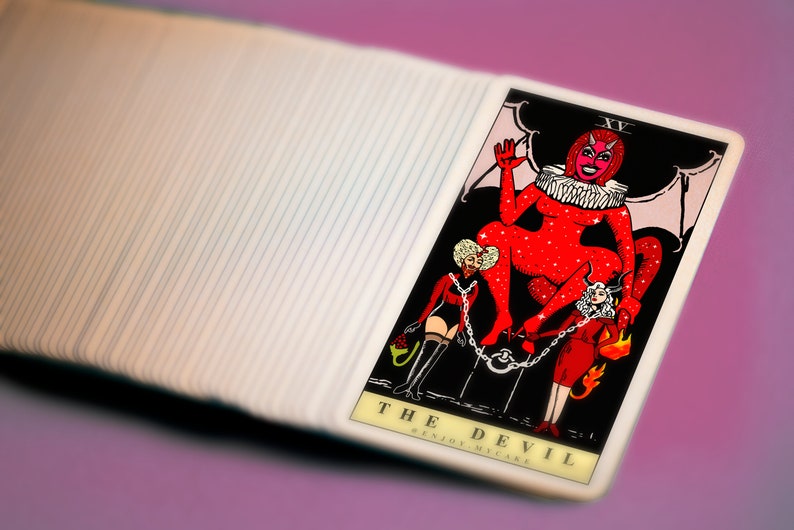 May include: The Devil tarot card features a red, sparkly figure with horns and wings, sitting on a throne. Two figures are chained to the throne, one in red and one in black. The card reads "THE DEVIL ENJOY  YOUR  MISTAKE".