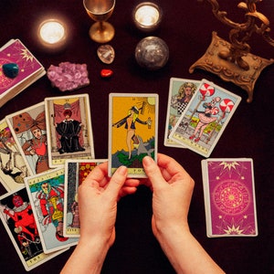 May include: A tarot card reading spread with a variety of cards laid out on a dark purple surface. The cards are surrounded by candles, crystals, and a gold candlestick. The person is holding the "The Fool" card, which depicts a figure standing on a cliff with a staff and a backpack.